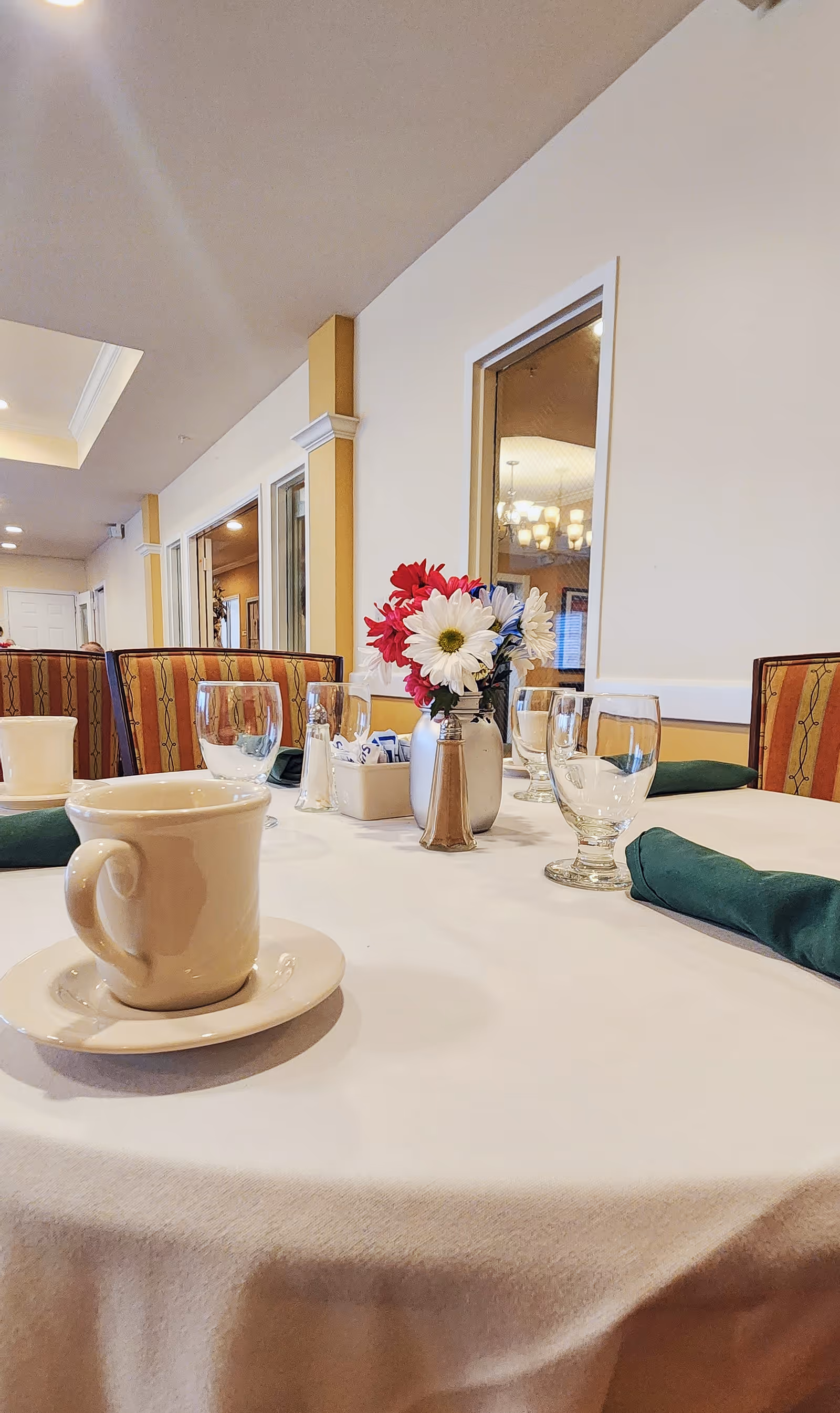 Set dining table in a communal dining room with a coffee cup, glassware, green napkins, and a small floral centerpiece.
