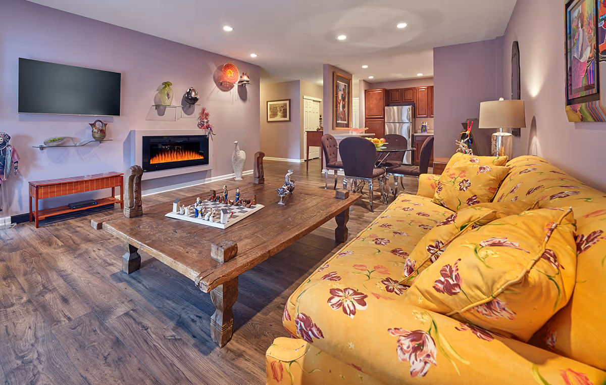 Bright living room with a yellow floral sofa, low rustic coffee table, wall-mounted TV and electric fireplace, with a dining area and kitchen visible in the background.
