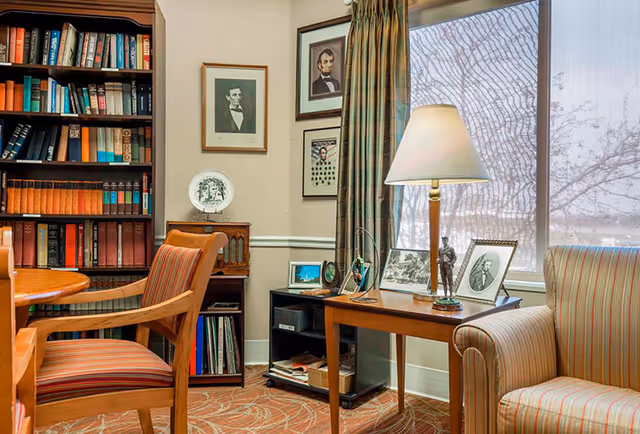 A cozy room with a wooden bookshelf filled with books, a wooden table with a striped cushioned chair, and a side table with a lamp and framed photos. The room has a large window with curtains and portraits on the wall.