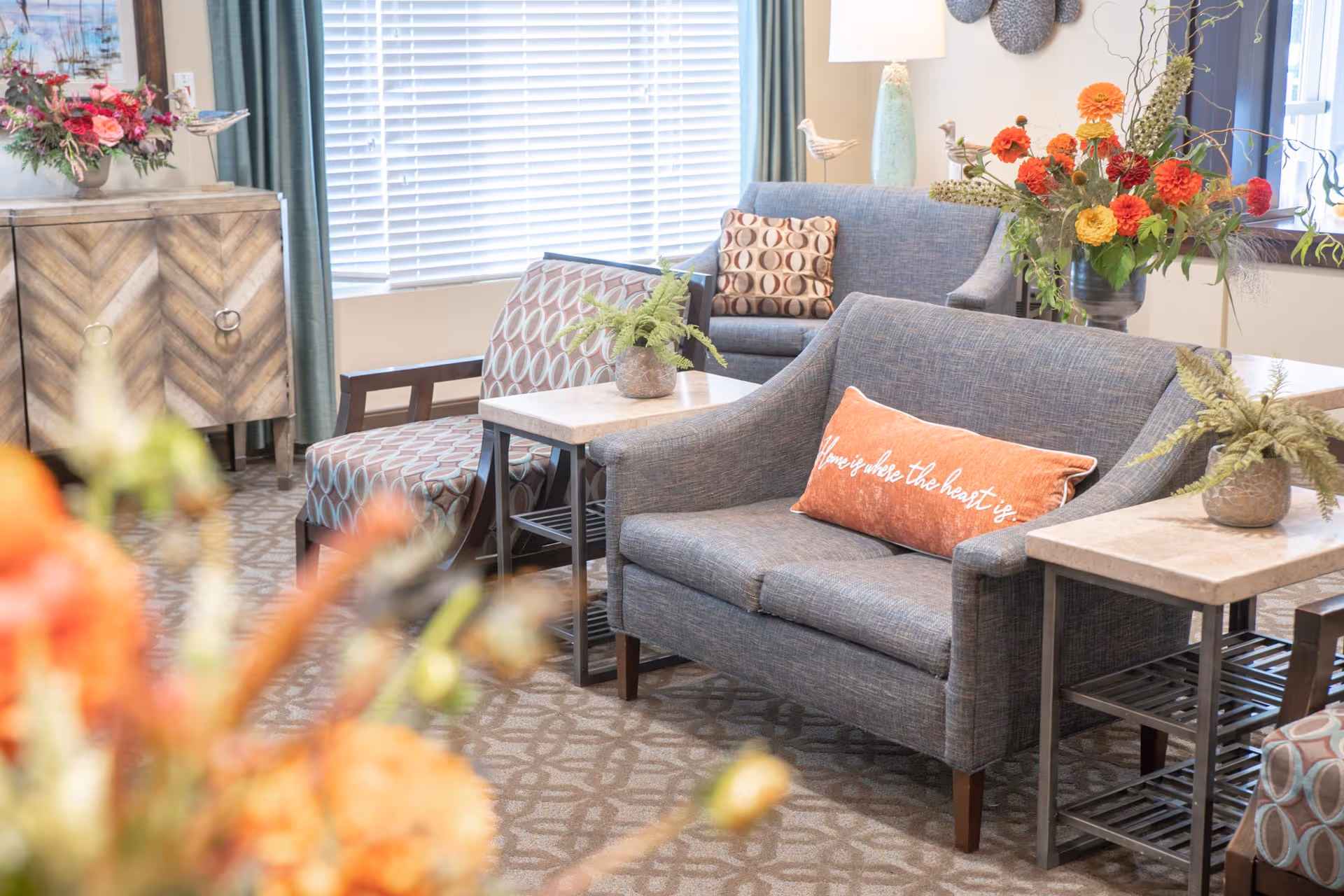 A cozy living room area with two gray upholstered loveseats, one with an orange pillow that reads 'Home is where the heart is.' There are patterned armchairs, small tables with potted plants, a wooden cabinet with a floral arrangement, and large windows with white blinds and green curtains.