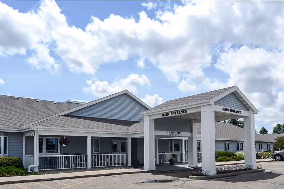 Exterior front of the Edgewood Minot senior living facility showing a covered main entrance and parking area under a partly cloudy sky.