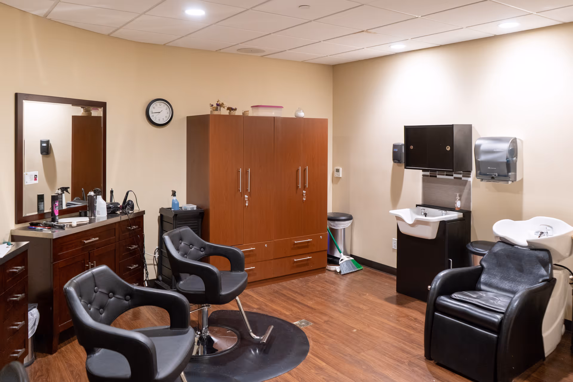 Interior of a salon room with two black salon chairs in front of a wooden cabinet and mirror. There are two white hair washing sinks with black chairs next to them. The room has wooden flooring, beige walls, and a clock on the wall.
