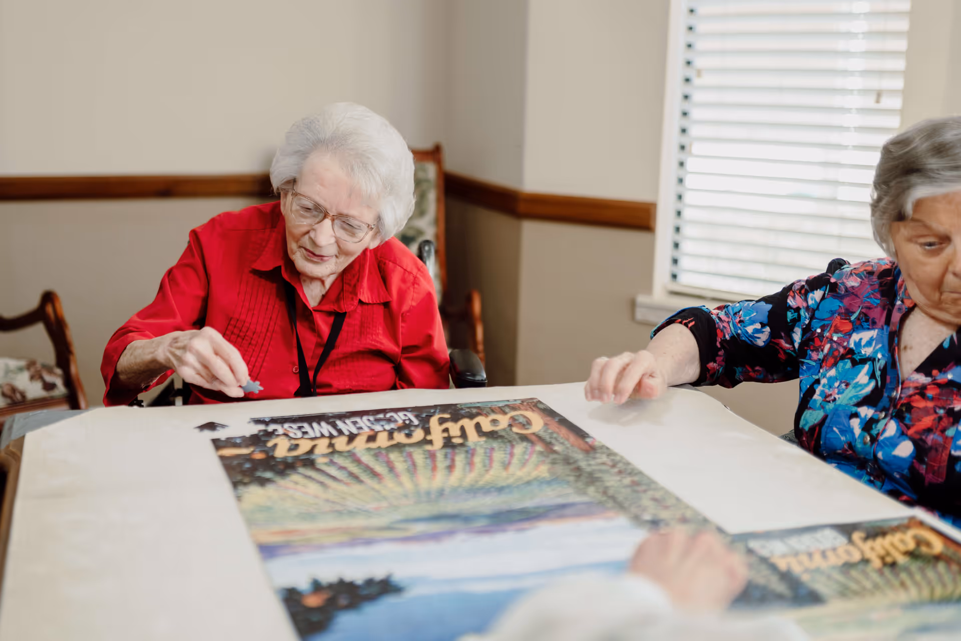 Two elderly women sitting at a table working on a large jigsaw puzzle together in a well-lit room with a window and wooden chairs.