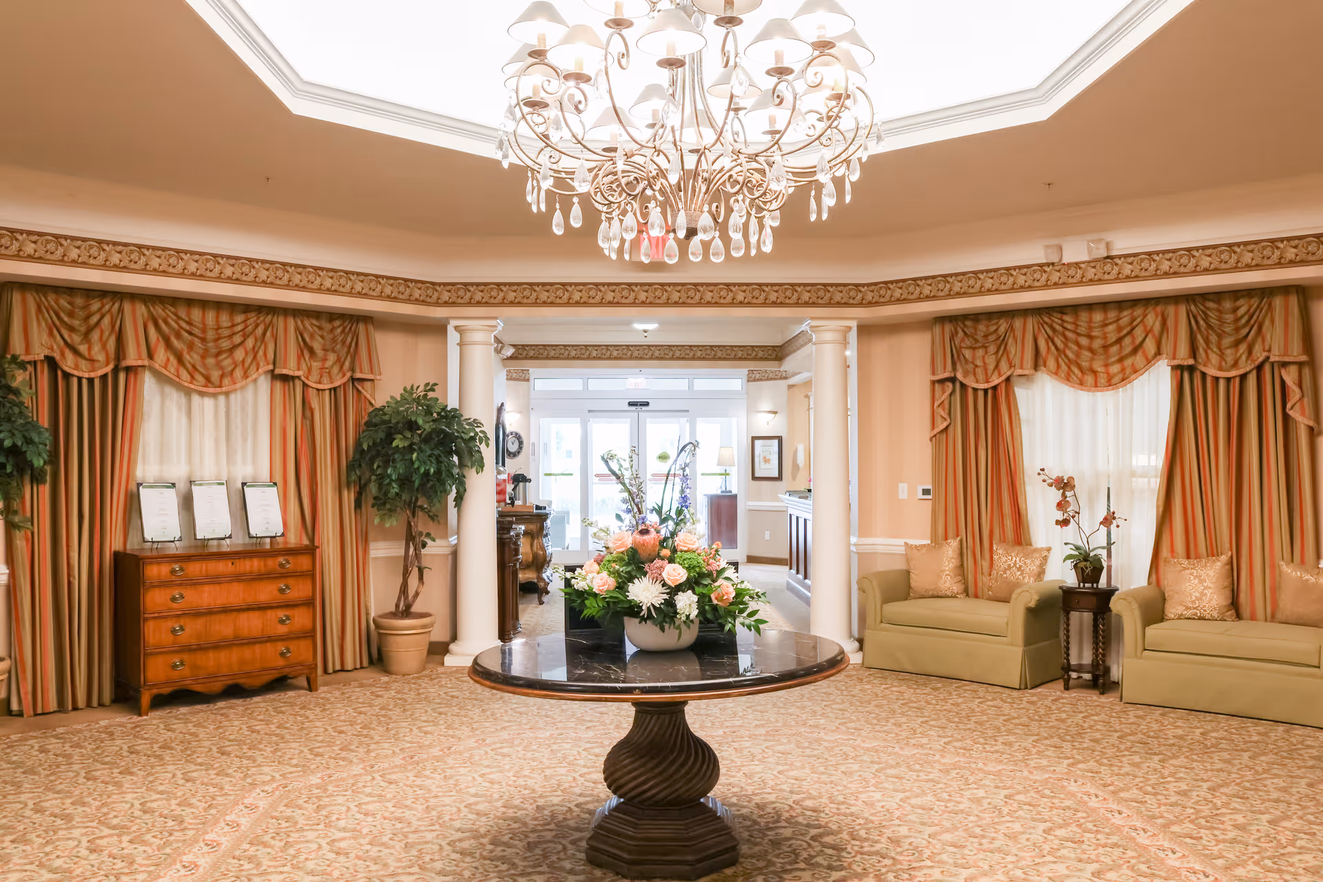 An elegant senior living facility lobby with a round table in the center holding a floral arrangement. The room features beige walls with decorative molding, two large windows with striped curtains, two green sofas with gold pillows, potted plants, a wooden chest of drawers, and a chandelier hanging from the ceiling.