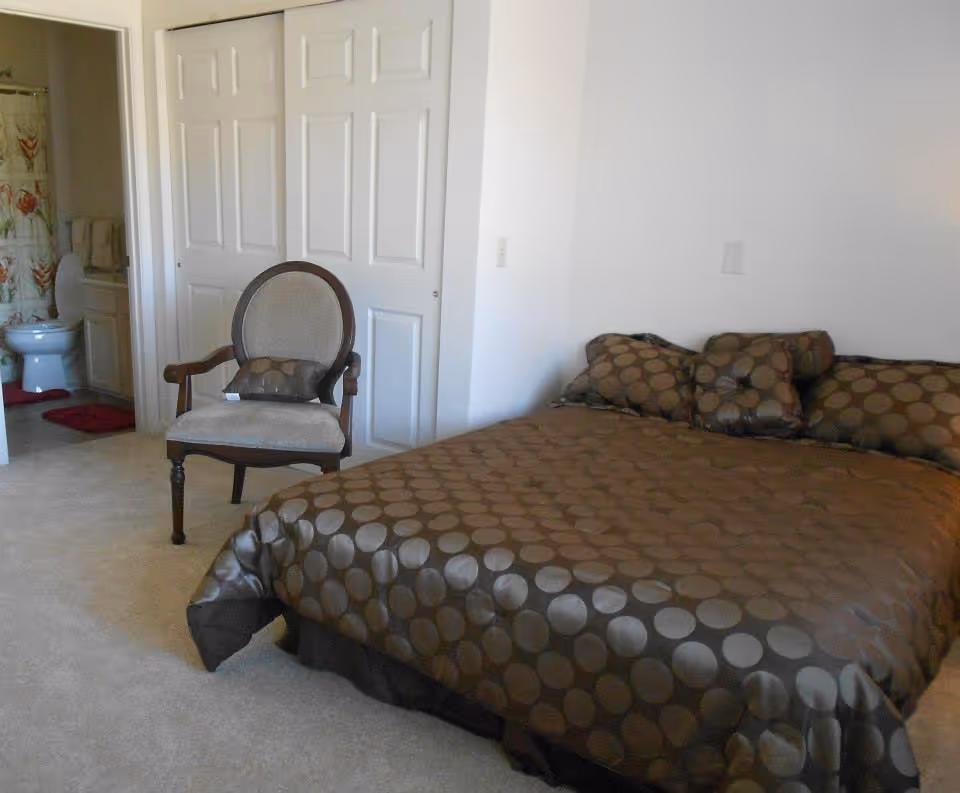 A bedroom with a large bed covered in a brown bedspread with circular patterns. There are matching pillows on the bed. Next to the bed is a wooden chair with a cushioned seat and backrest, also with a small pillow. In the background, there is a bathroom visible with a toilet, sink, and a shower curtain with a floral design. The room has light-colored carpet and white walls.