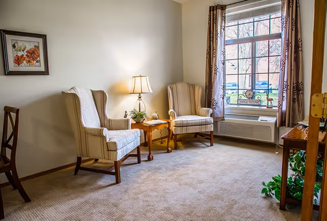 A cozy sitting area with two upholstered armchairs, a side table with a lamp, and a window with curtains.