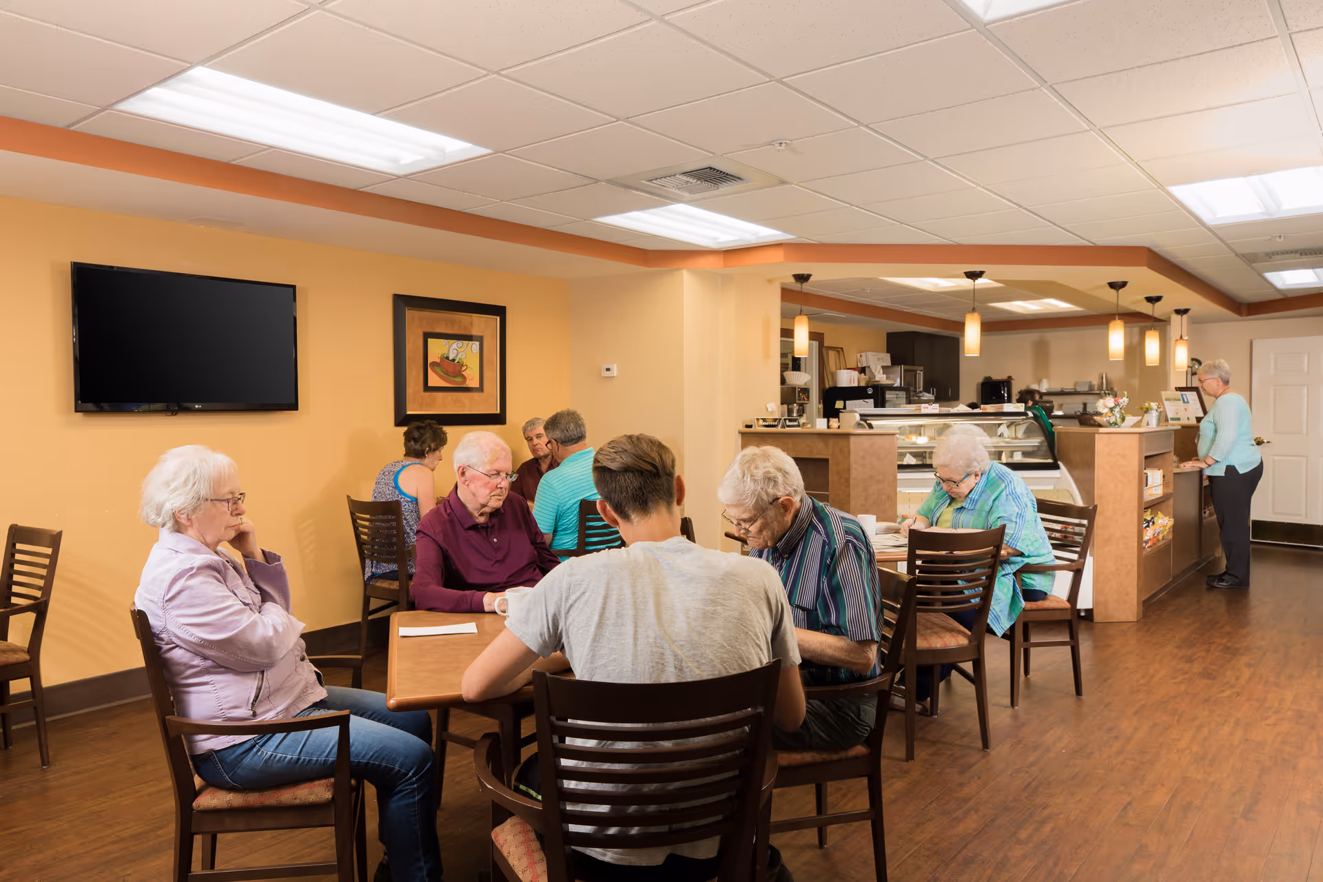 A group of elderly people sitting and socializing at tables in a warm, well-lit common area with wooden floors and beige walls. There is a flat-screen TV mounted on the wall, a framed picture, and a counter area in the background where a woman is standing. Pendant lights hang from the ceiling above the counter.