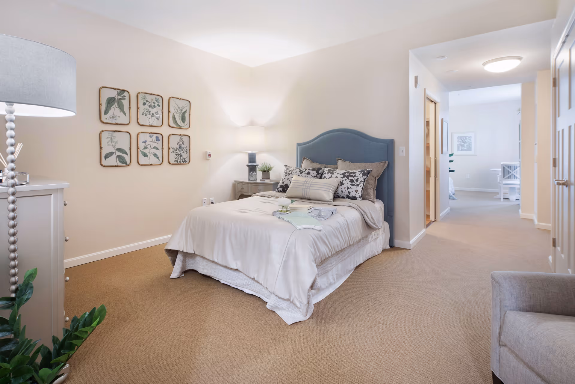 A neatly made bed with a blue upholstered headboard and multiple pillows in a softly lit bedroom. The room features beige walls and carpet, a bedside table with a lamp and plant, framed botanical prints on the wall, and a hallway leading to another room with a table and chairs visible in the distance.