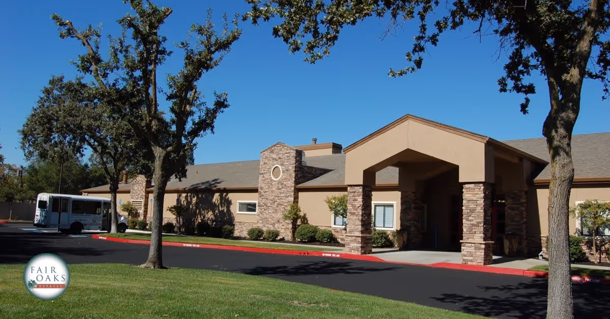 Exterior view of Fair Oaks Estates building with stone and beige stucco facade, a covered entrance supported by stone columns, surrounded by trees and a parking area with a shuttle bus parked on the left side under a clear blue sky.