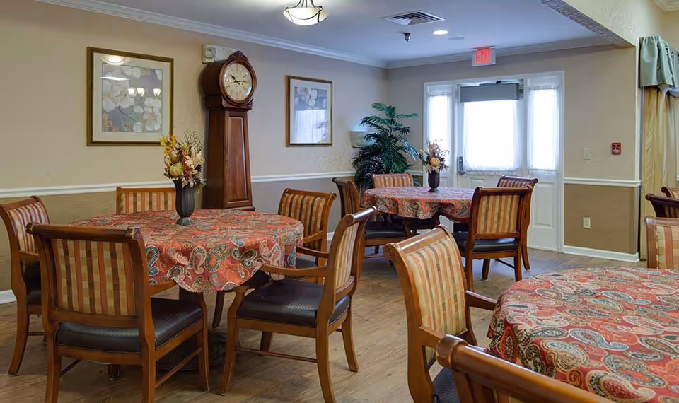 A dining room with round tables covered in red patterned tablecloths, each surrounded by wooden chairs with cushioned seats and backs. The room features a wooden grandfather clock, framed floral artwork on the walls, a potted plant near a door with windows covered by sheer curtains, and a wooden floor.