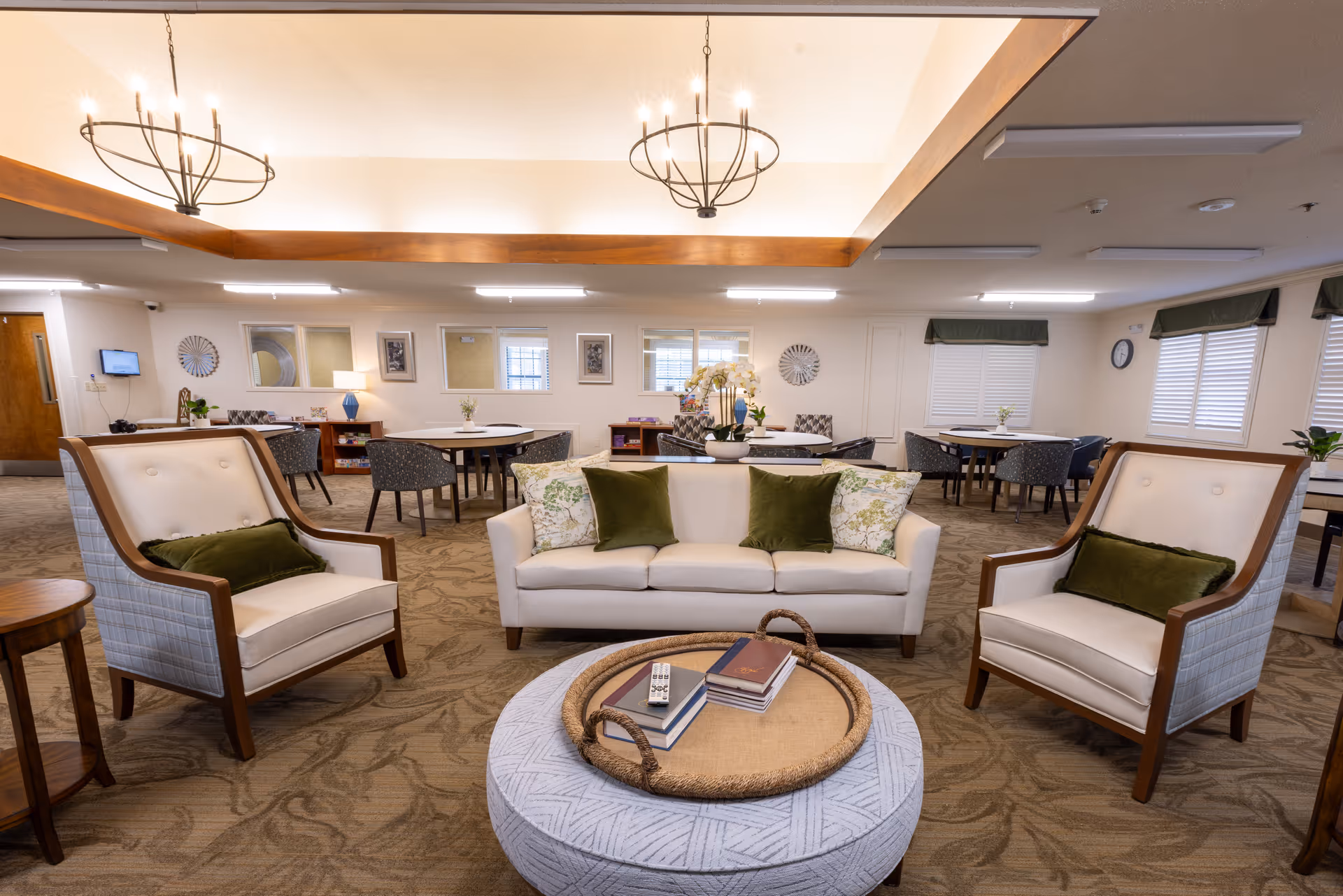 A spacious and well-lit living room area in a senior living facility with a white sofa adorned with green and floral pillows, two matching armchairs with green cushions, a round ottoman with a woven tray holding books and a remote control, and several round tables with chairs in the background. The room features beige carpeting, decorative wall clocks, framed pictures, and large windows with white blinds and green valances.