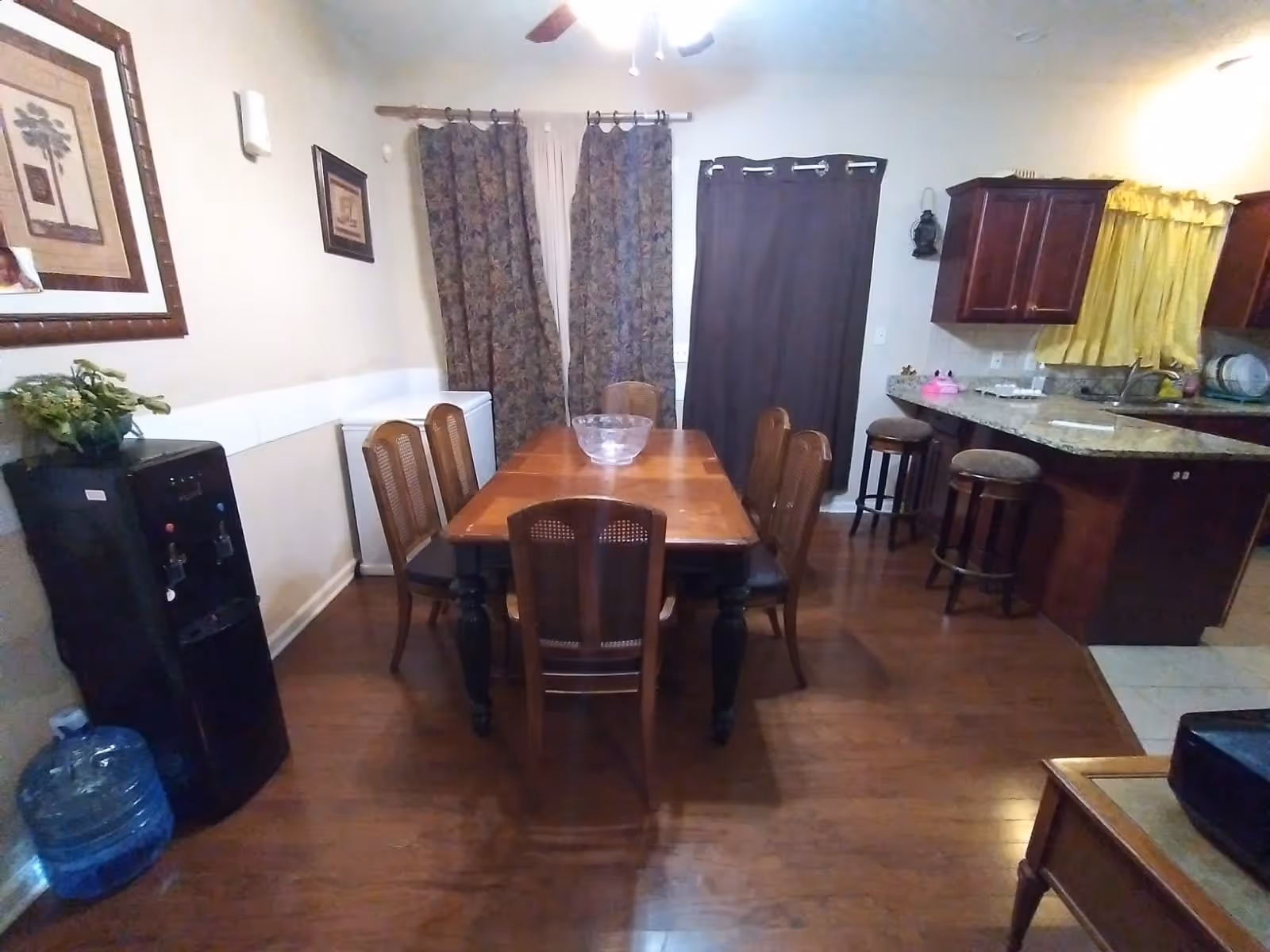 Interior view of a dining area with a wooden dining table surrounded by six chairs. The room has hardwood flooring, a water dispenser with a large water bottle on the floor, framed artwork on the walls, and windows covered with patterned and solid curtains. To the right, there is a kitchen area with granite countertops, wooden cabinets, and two bar stools.