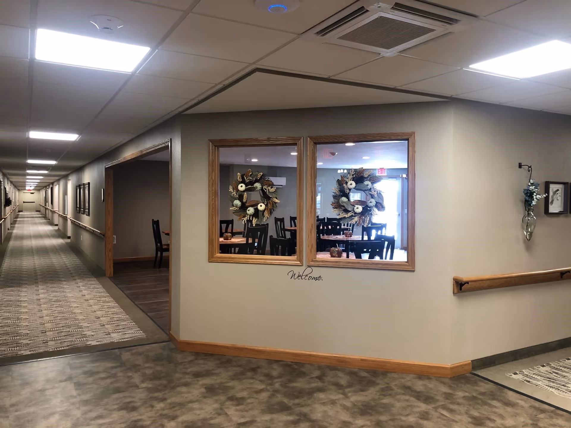 Well-lit assisted living interior hallway with handrails and a windowed dining room decorated with wreaths and a "Welcome" sign.