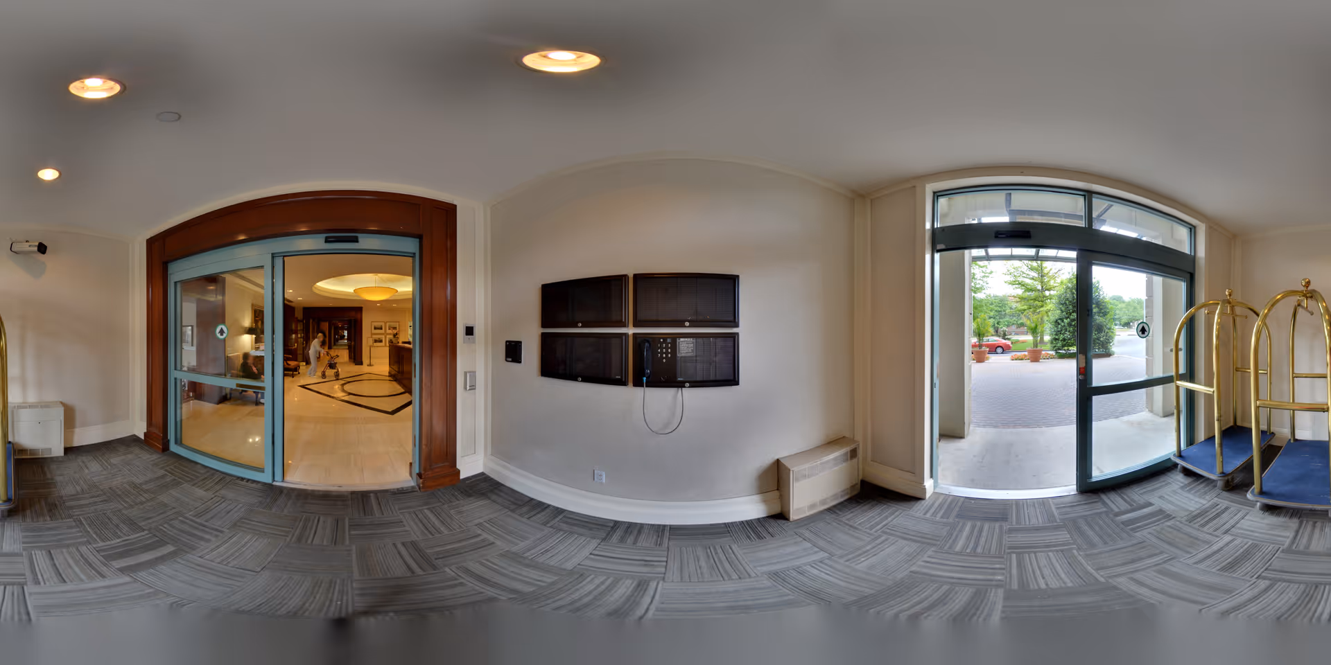 Entrance area of a senior living facility with automatic sliding glass doors leading inside to a lobby where a person is visible. The area has carpeted flooring with a patterned design, a wall-mounted intercom or communication system, and luggage carts near the exit door leading outside to a paved area with greenery and parked cars.