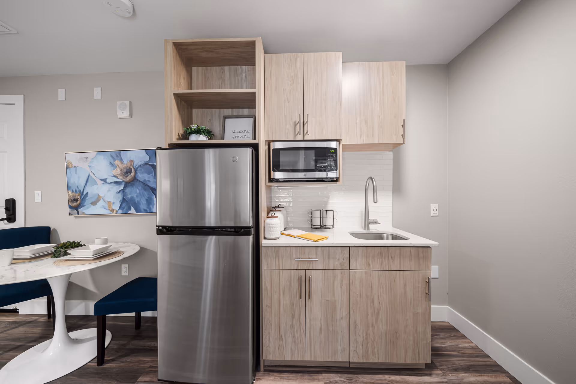 Modern kitchenette with light wood cabinets, a stainless steel refrigerator, a microwave, and a sink. To the left, there is a small dining area with a white round table, blue chairs, and a wall painting featuring large blue flowers.
