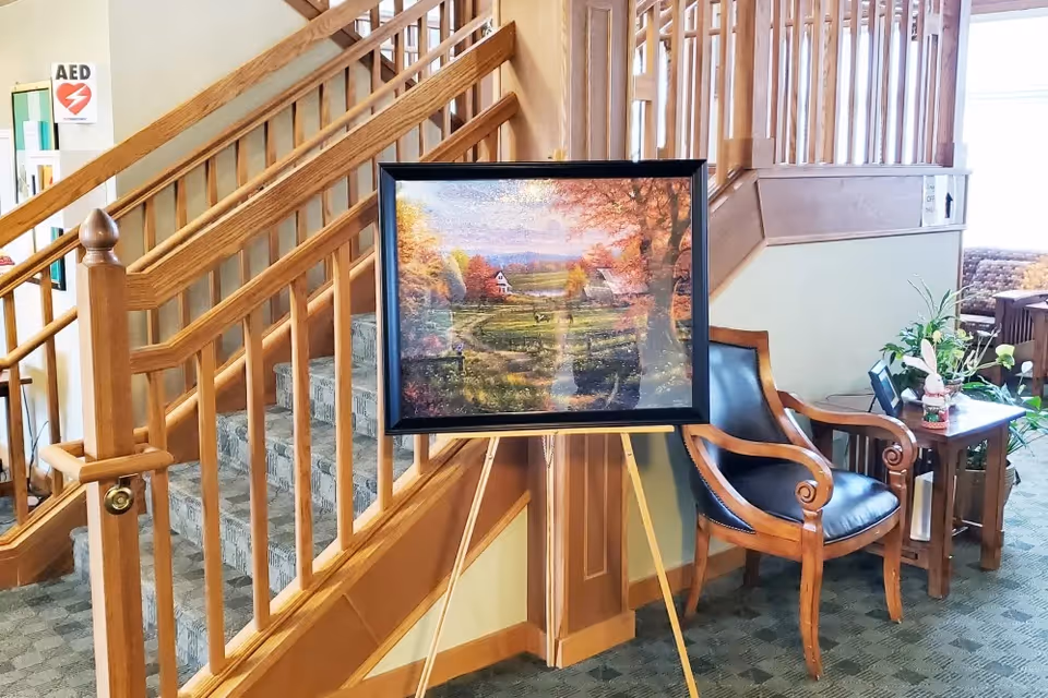 Interior common area showing a wooden staircase, an easel with a framed landscape painting, and a leather armchair beside a small table with plants.