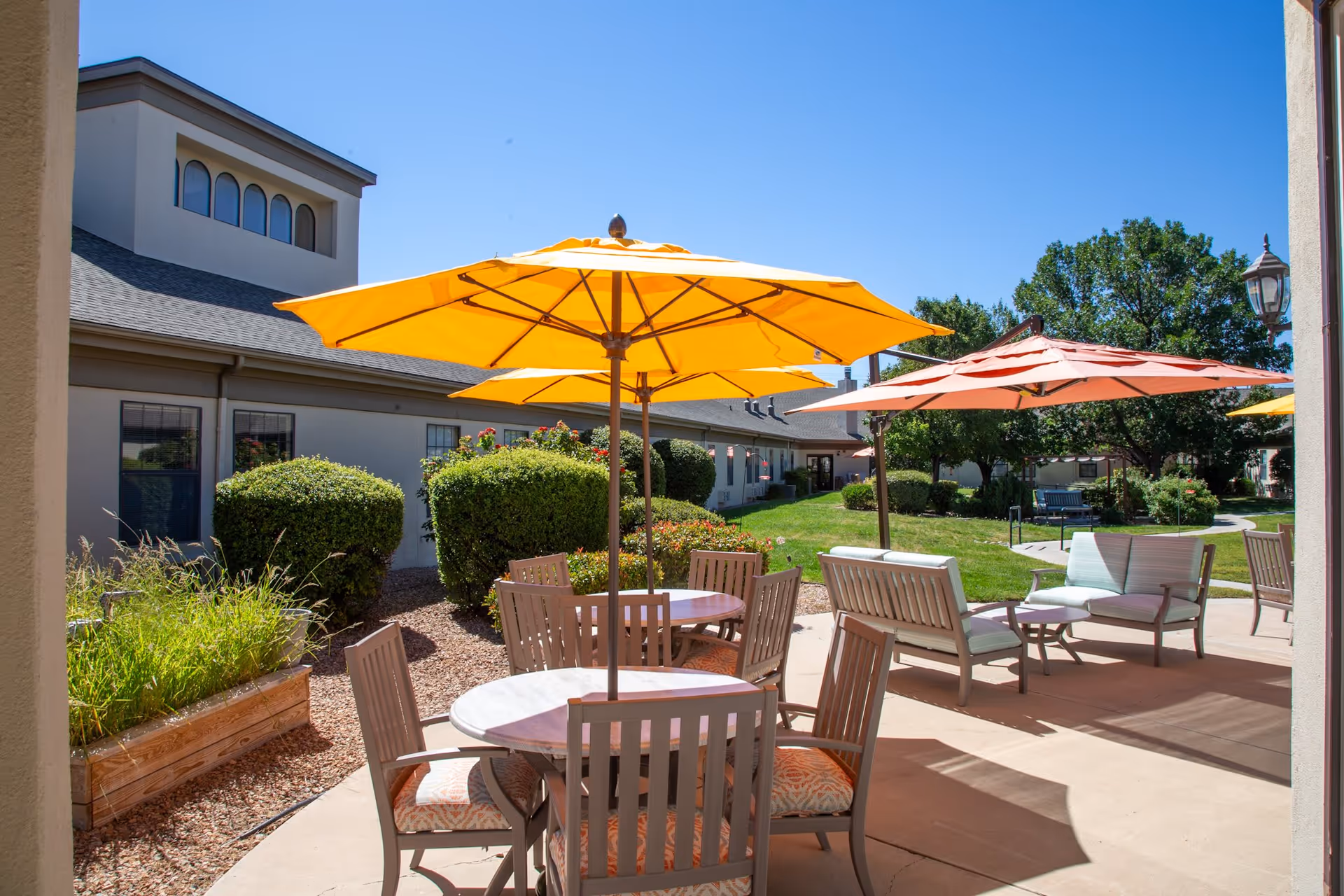 Outdoor patio area with round tables, chairs, and large yellow and orange umbrellas providing shade. The patio is adjacent to a building with bushes and greenery surrounding the area under a clear blue sky.
