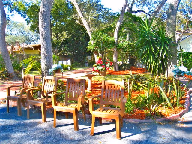 Outdoor garden area with four wooden chairs arranged in a row on a paved surface. The background features trees, various plants, flowers, and a mulched garden bed under bright sunlight.