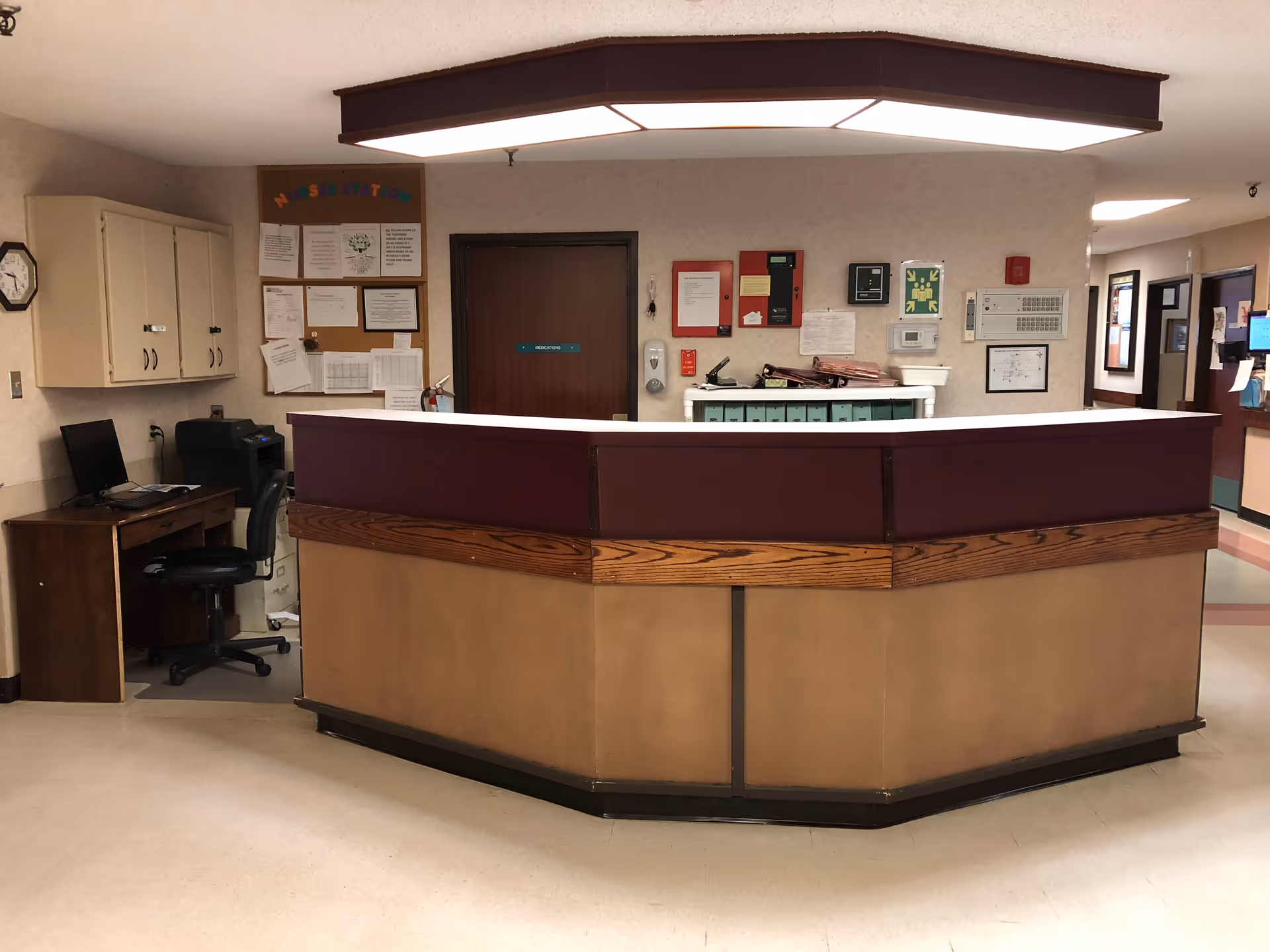 A reception/nurse station desk in the hallway of a senior care facility with bulletin boards, office workspace, and corridor doors.