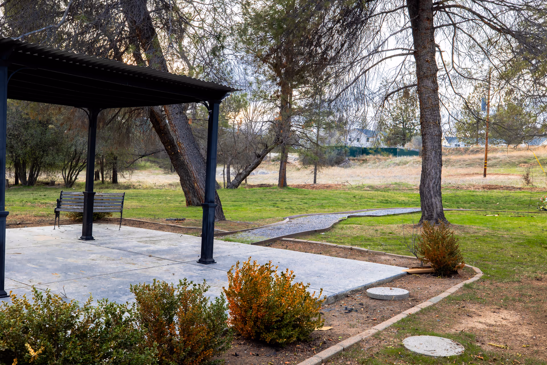Concrete patio with a metal pergola and bench overlooking a grassy yard with trees and a winding gravel path.