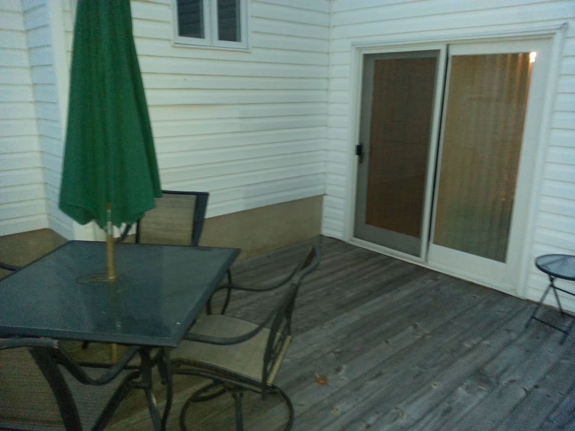Outdoor wooden deck area with a glass-top table, four chairs, and a closed green patio umbrella. The deck is adjacent to a white siding building with a sliding glass door and a window.