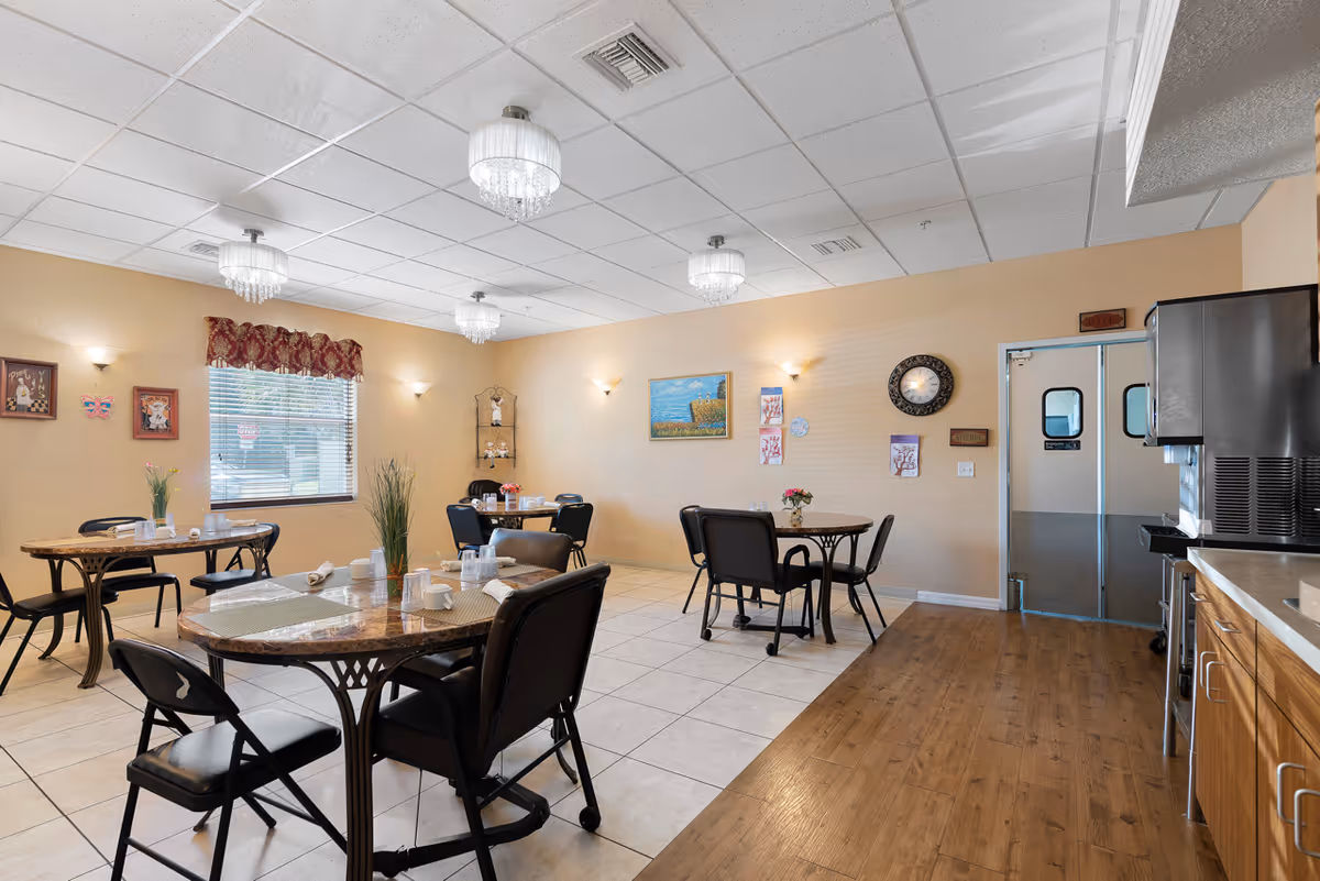 A dining room in an assisted living facility with several round tables and black chairs. The tables are set with placemats, napkins, cups, and small flower arrangements. The room has beige walls, a window with a red valance, wall-mounted lights, and ceiling chandeliers. There is a clock and artwork on the walls, and a kitchen area with cabinets and a beverage dispenser is visible on the right side.