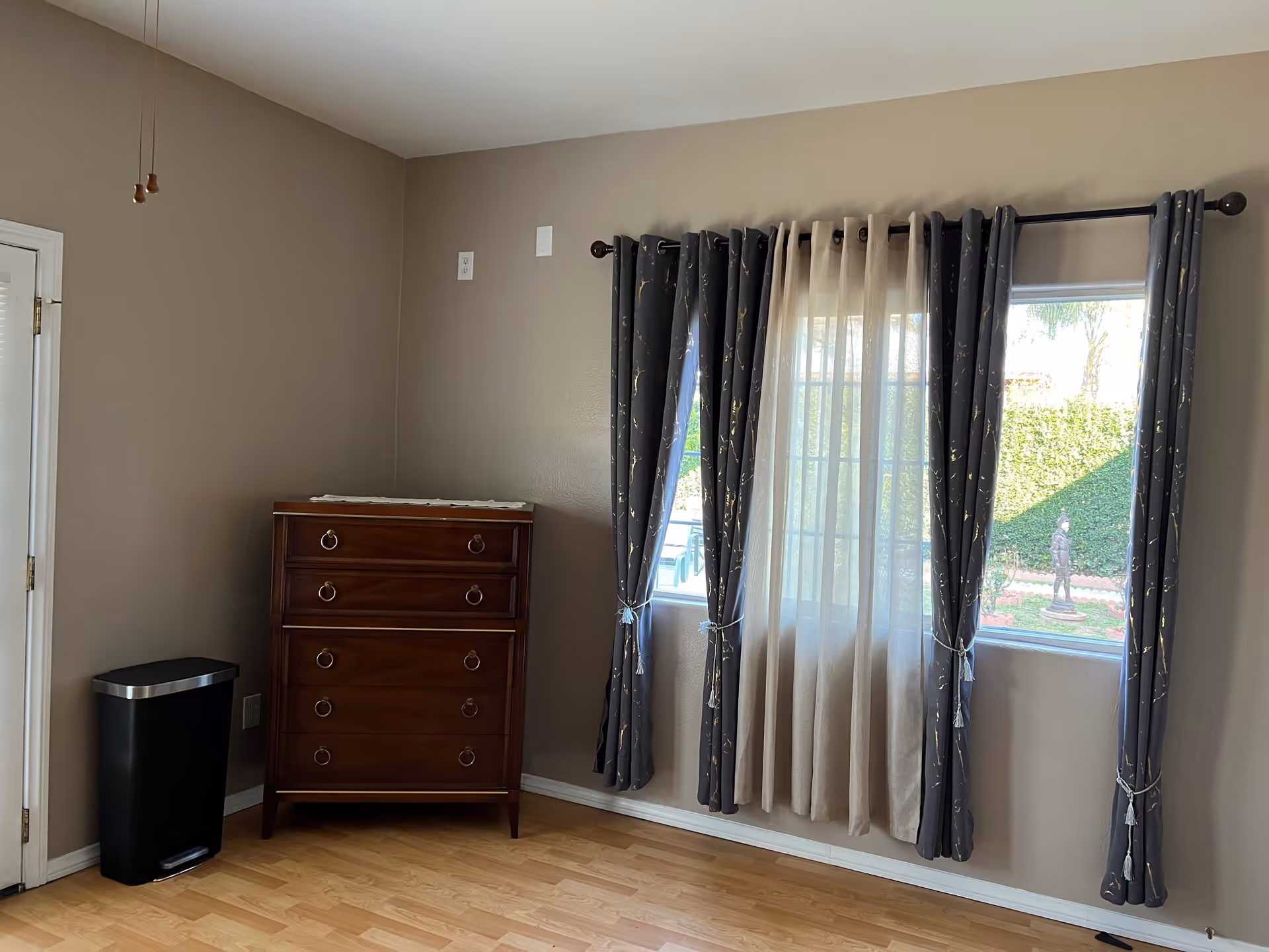 A corner of a room with beige walls and light wood flooring. There is a wooden chest of drawers with four drawers and ring handles, next to a black trash bin with a silver lid. Two windows with dark blue curtains and sheer beige drapes let in natural light, showing a garden with a statue outside.