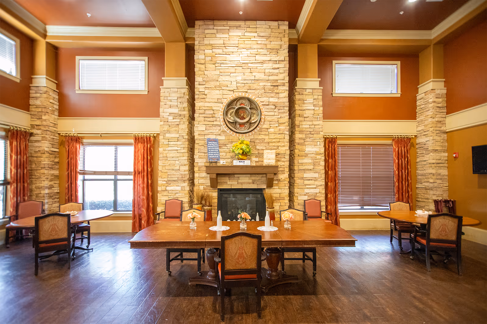 A warm and inviting dining room with a large wooden table in the center, surrounded by chairs. The room features a stone fireplace with decorative items on the mantel, tall windows with red curtains, and wooden flooring. Additional smaller tables and chairs are placed near the windows.