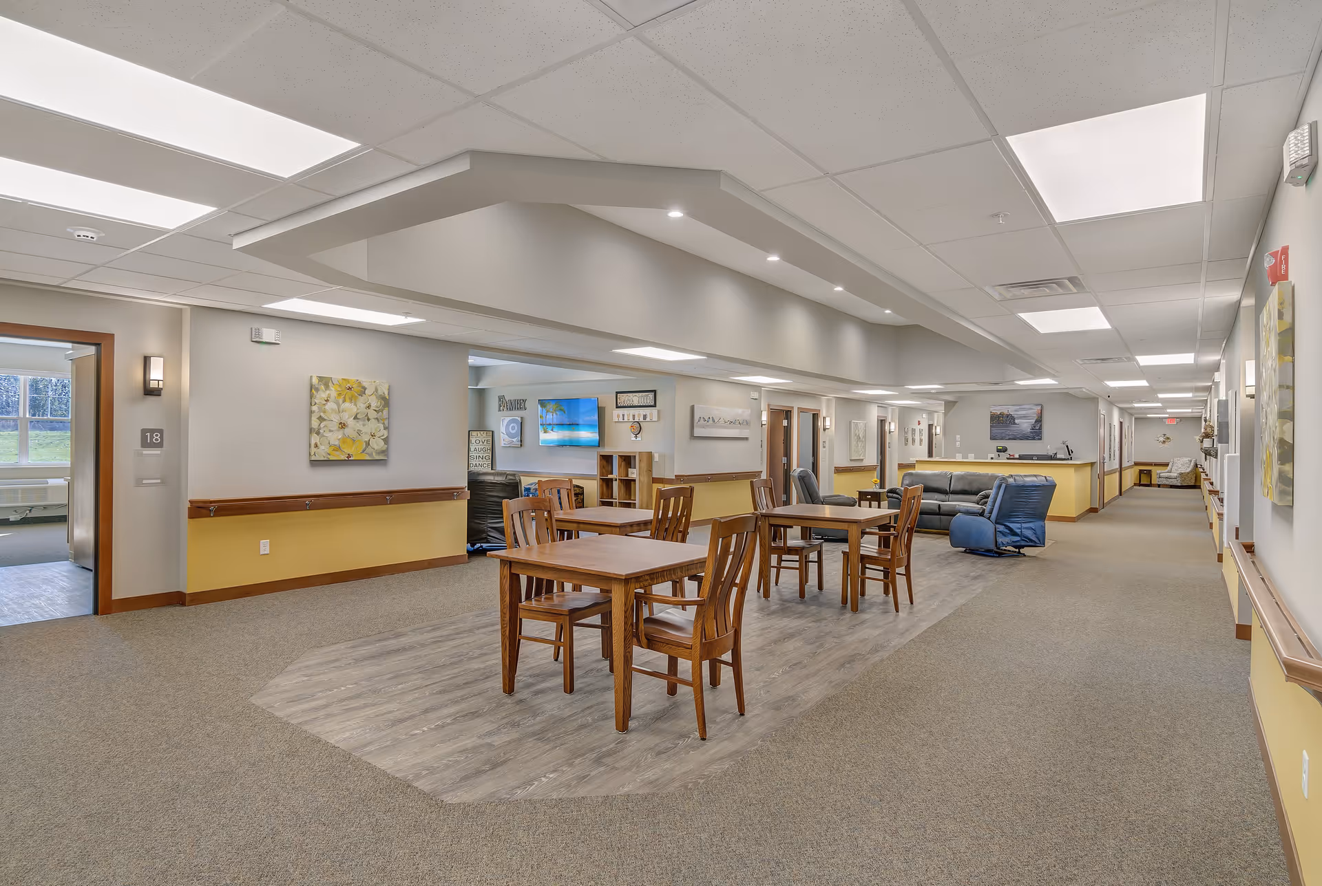 A spacious and well-lit common area in a senior living facility featuring several wooden tables with chairs on a wood-patterned floor section, surrounded by carpeted flooring. The area includes a seating section with a black sofa and a blue recliner chair. The walls are decorated with paintings and there is a television mounted on the wall. The hallway extends to the right with doors leading to other rooms.