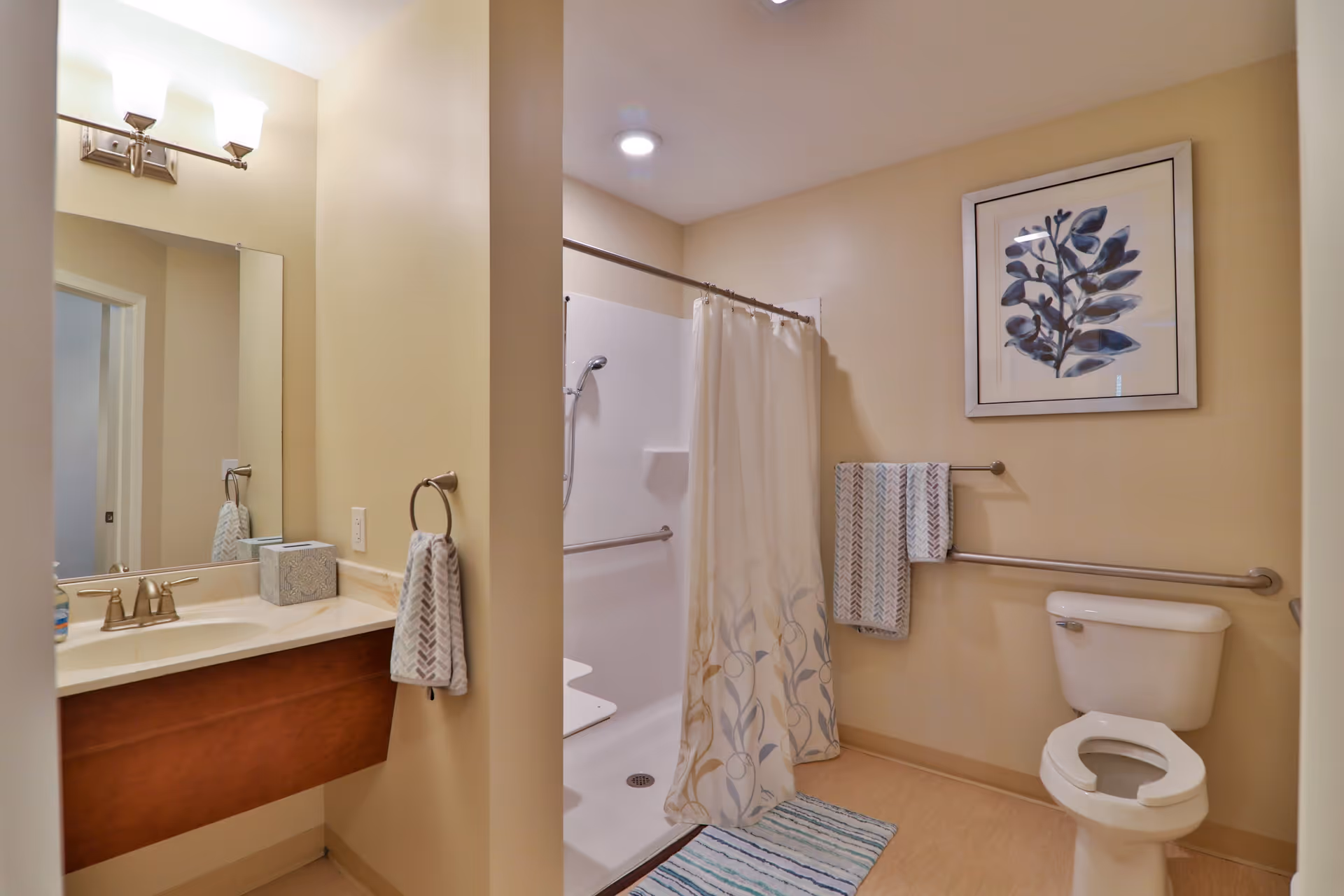 A senior-friendly bathroom with beige walls featuring a walk-in shower with a floral shower curtain, a white toilet with a raised seat, grab bars on the walls, a towel rack with two patterned towels, a framed botanical artwork, and a vanity with a sink, mirror, and light fixture above.
