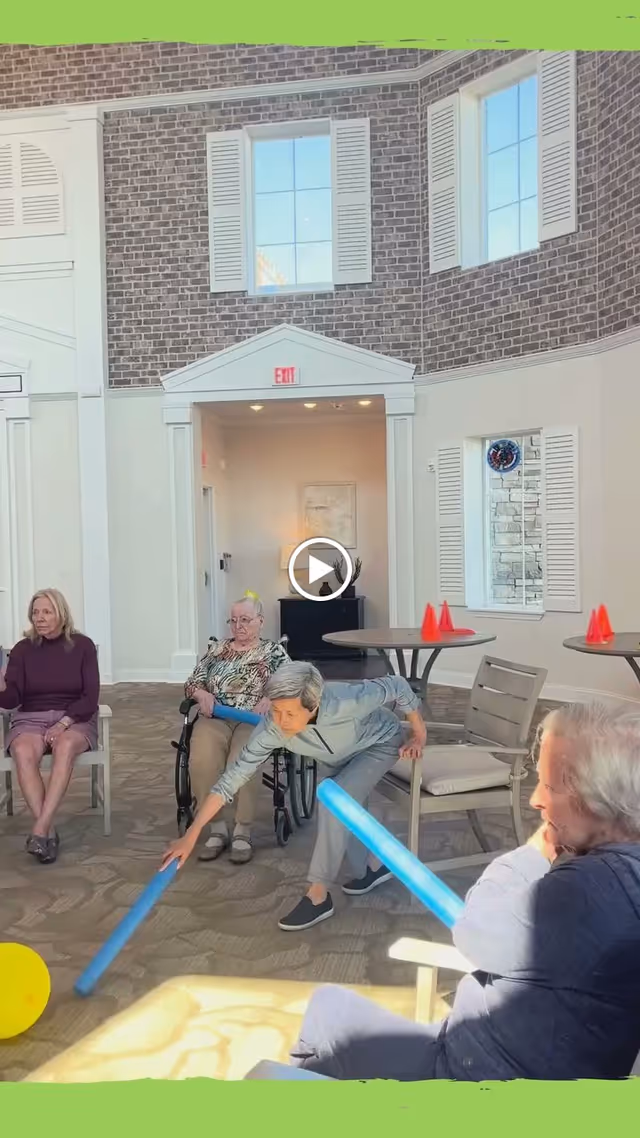 A group of elderly women in a bright, spacious room with large windows and brick accent walls. One woman in a wheelchair and others seated in chairs are engaged in a light physical activity using blue foam sticks and a yellow ball. The room has round tables with orange cones on them and an exit sign above a doorway.