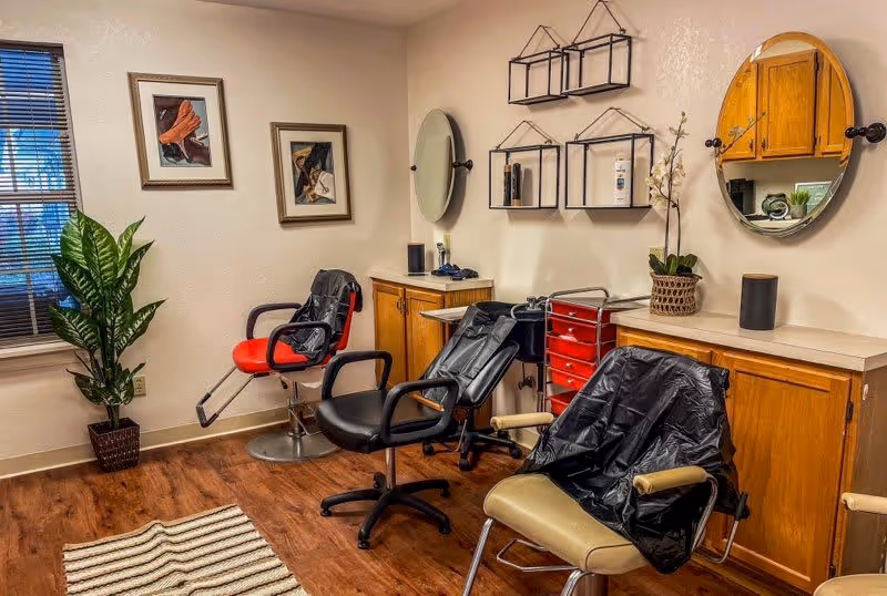 Interior of a salon area with three styling chairs, two round mirrors on the wall, wooden cabinets, a red rolling cart with drawers, a potted plant, two framed pictures on the wall, and a window with blinds.