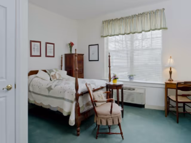 A cozy bedroom with a wooden four-poster bed covered in a quilted bedspread. There is a wooden chair with a cushion in front of the bed, a small round table with a flower pot, a wooden dresser with a vase of flowers, and a wooden desk with a lamp and chair near a large window with blinds and a valance.