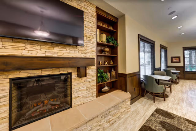 Cozy common room with a stone fireplace, mounted TV, built-in shelving and seating by windows.