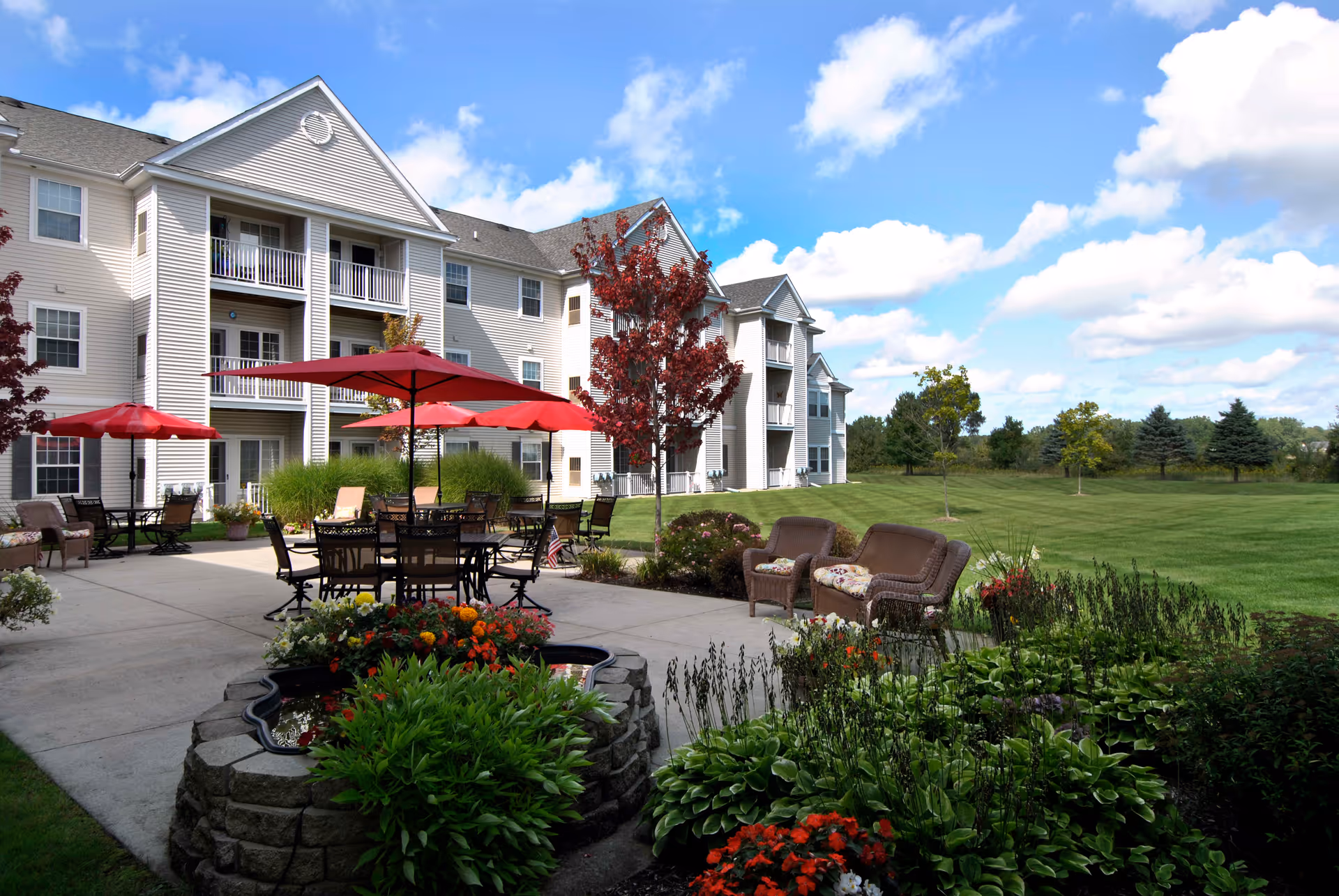 Outdoor patio area at a retirement community with tables and chairs under red umbrellas, surrounded by landscaped gardens and green lawn, with a multi-story residential building in the background under a partly cloudy blue sky.