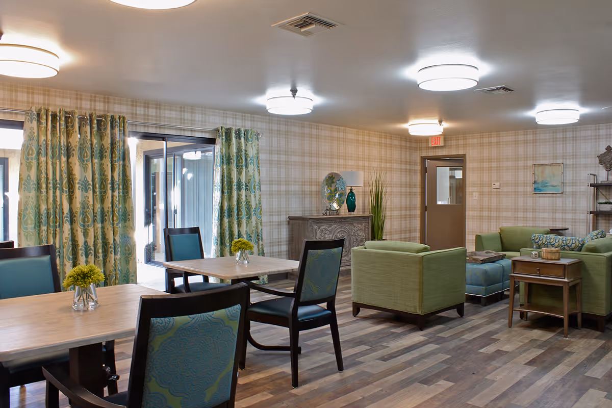 A well-lit common area in a senior living facility featuring wooden tables with green cushioned chairs, green armchairs, a green sofa, and a blue ottoman. The room has patterned curtains, plaid wallpaper, wooden flooring, and ceiling lights. There is a decorative cabinet with a lamp and vase, a small side table, and a door with an exit sign above it.