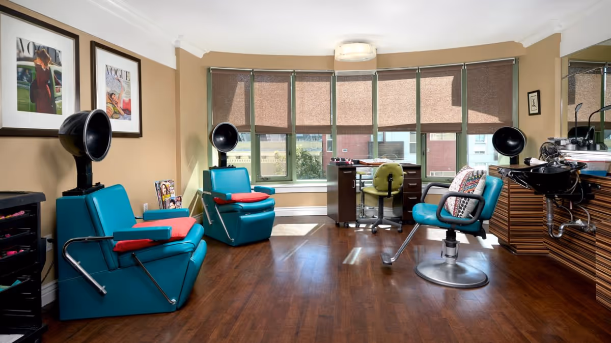 Bright salon-style interior with teal styling chairs, hooded hair dryers, a wash basin and a desk in front of a large bay window.