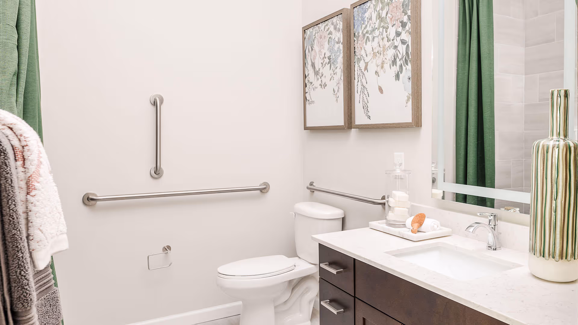 Well-lit bathroom featuring a toilet, sink vanity with countertop and mirror, stainless grab bars, framed artwork, and a green shower curtain.