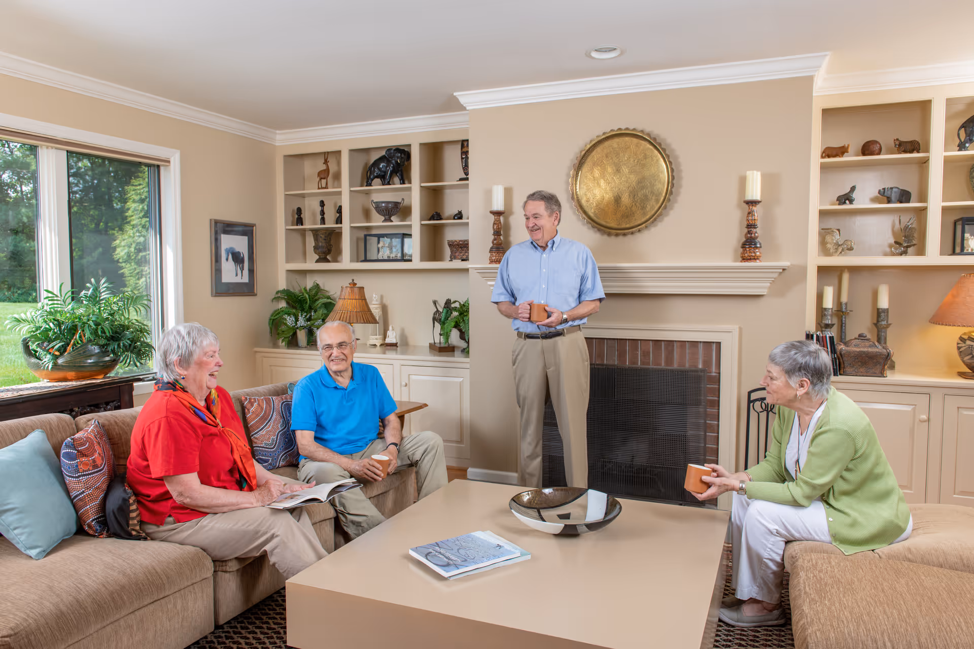 Four elderly people socializing in a bright living room with a fireplace, built-in shelves, and a large coffee table.