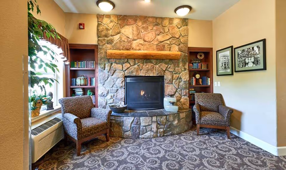 Cozy seating area with a stone fireplace flanked by built-in bookshelves, two upholstered armchairs, a window and decorative plants.