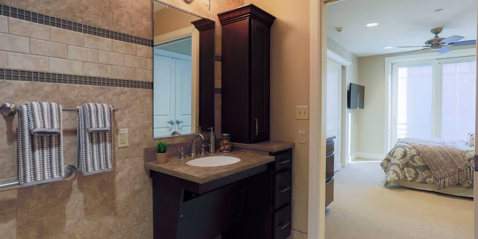Bathroom vanity with sink, towel rack on tiled wall, and a view into a bedroom with a bed and TV.