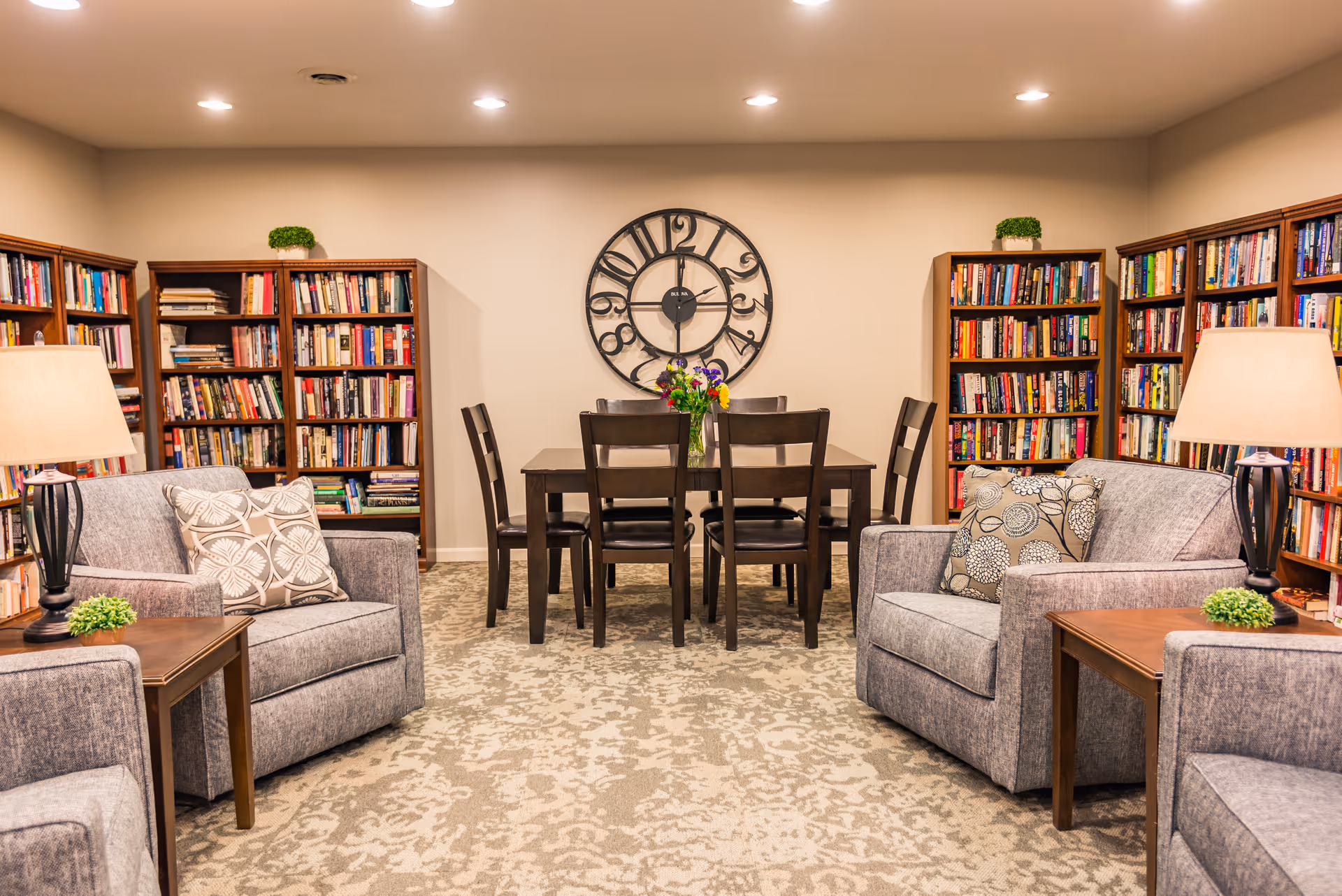 Cozy communal library lounge with bookshelves along the walls, upholstered armchairs and side tables, and a central table with chairs beneath a large wall clock.