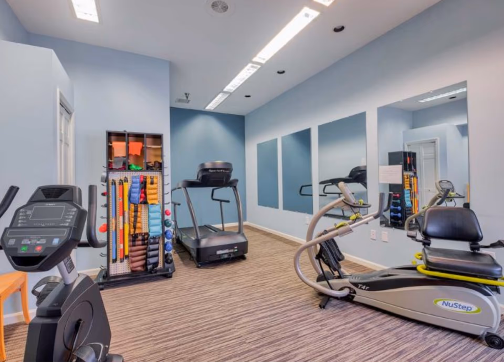 A small fitness room with exercise equipment including a stationary bike, treadmill, and a NuStep recumbent cross trainer. There is a rack holding various exercise bands and small weights. The room has light blue walls, three large mirrors on one wall, and overhead fluorescent lighting.