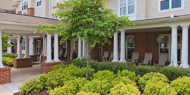 Outdoor view of Hampshire Village showing a covered porch area with white columns and several rocking chairs. In front of the porch, there is a well-maintained garden with green shrubs and a small tree.