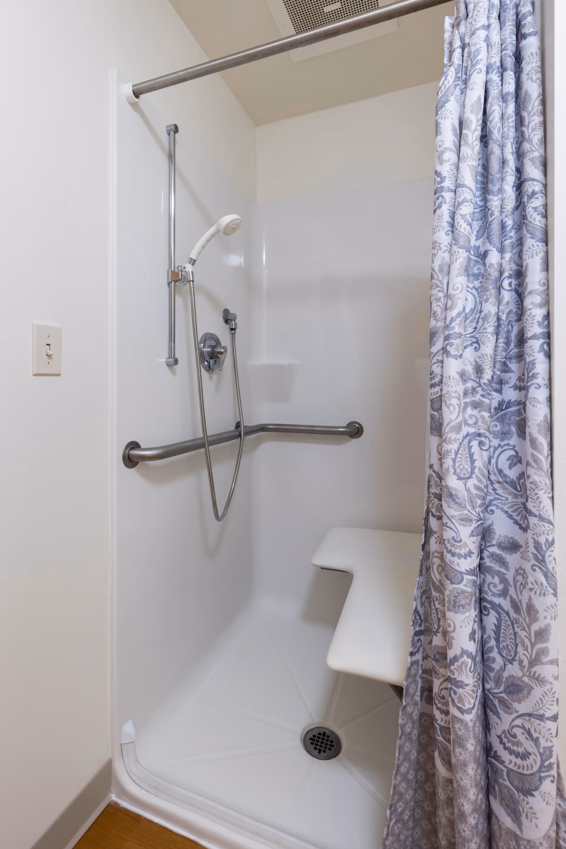 A white shower stall with a handheld showerhead attached to a vertical sliding bar, a horizontal grab bar, and a built-in white shower seat. The shower curtain has a blue and white floral pattern.