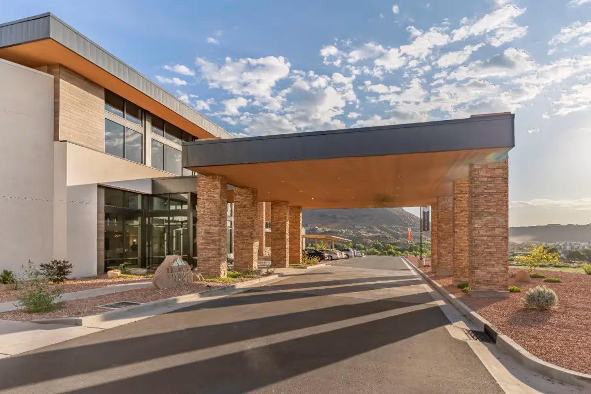 Front entrance canopy of a modern senior living facility with stone columns, a driveway, and desert landscaping under a blue sky.
