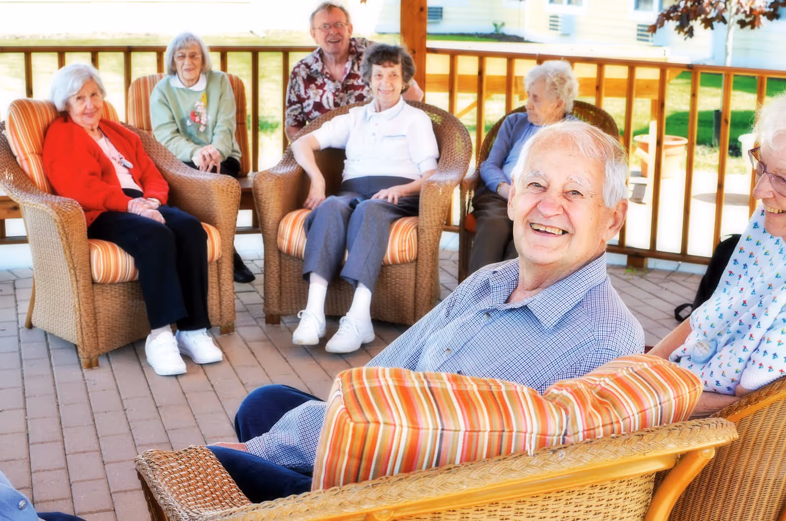 A group of elderly people sitting and smiling on a covered outdoor patio with wicker chairs and striped cushions, enjoying a sunny day.
