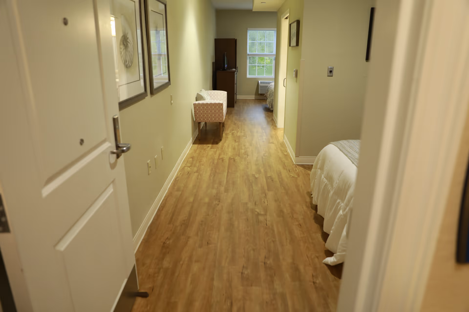 Hallway view into a senior living apartment with wood floors, seating, and a bed visible at the far end.