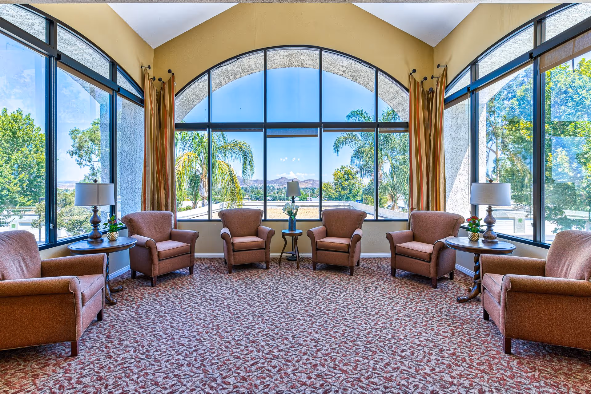 A bright and spacious sitting area with six brown upholstered armchairs arranged in a semi-circle around two round wooden side tables with lamps and small plants. Large floor-to-ceiling windows with an arched top provide a scenic view of palm trees, greenery, and distant hills under a clear blue sky. The room has patterned carpet flooring and light-colored walls.