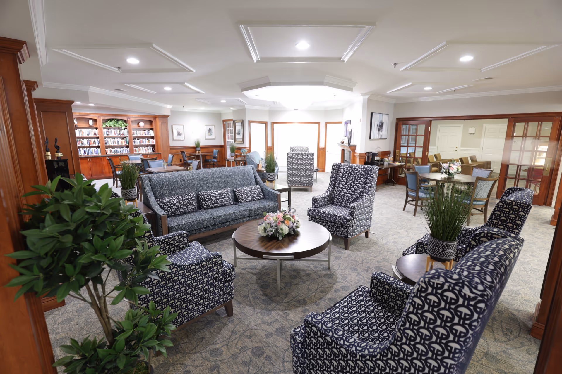 A spacious and well-lit senior living facility common area with patterned armchairs and a sofa arranged around a round wooden coffee table with a floral centerpiece. The room features carpeted floors, wood-paneled walls, potted plants, and a bookshelf filled with books. There are additional tables and chairs in the background, along with large windows and glass-paneled doors.
